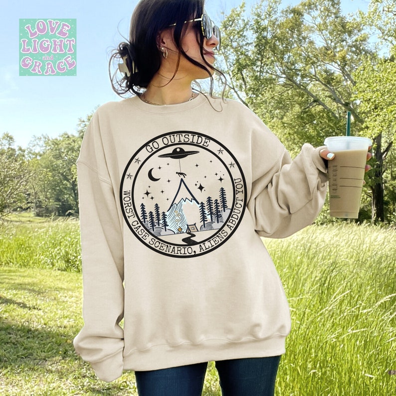 May include: A beige sweatshirt with a black and white graphic print of a mountain, a UFO, and stars. The text on the graphic reads "Go Outside Worst Case Scenario, Aliens Abduct You."