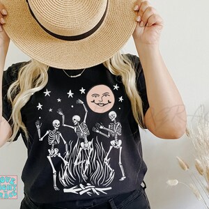 May include: Black t-shirt with a white graphic of three skeletons dancing around a bonfire with a smiling moon and stars in the background.