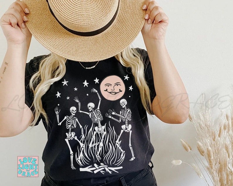 May include: Black t-shirt with a white graphic of three skeletons dancing around a bonfire with a smiling moon and stars in the background.
