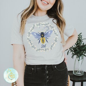 May include: A gray t-shirt with a floral wreath design featuring a bee with wings, a crescent moon, and stars. The text "Still." is printed below the bee.