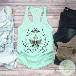 May include: A mint green tank top featuring a black and gold moth design, crystals, and feathers. The design is encircled by a black floral wreath.