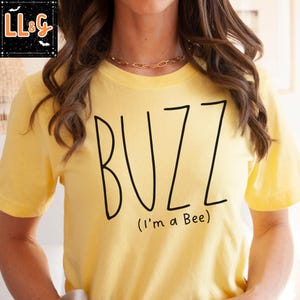 May include: A yellow t-shirt with the word "BUZZ" in large black letters. Below the word "BUZZ" is the phrase "(I'm a Bee)" in smaller black letters. The shirt is a solid yellow color.