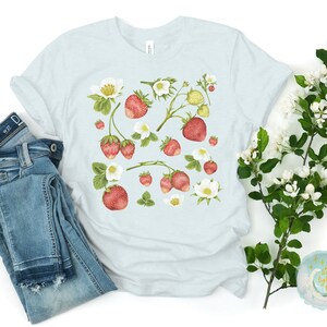 May include: A light blue t-shirt featuring a watercolor design of strawberries, blossoms, and leaves. The strawberries are red with green leaves and white blossoms. A pair of blue jeans is folded next to the shirt.