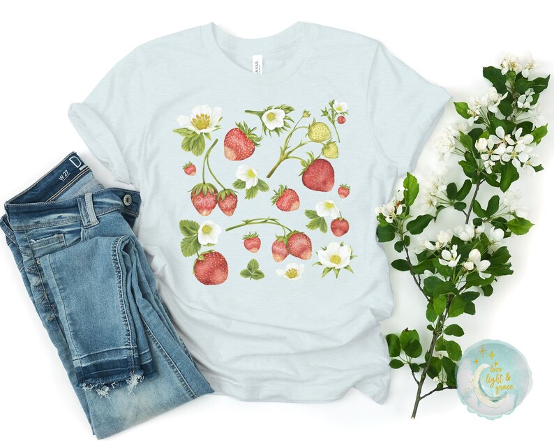 May include: A light blue t-shirt featuring a watercolor design of strawberries, blossoms, and leaves. The strawberries are red with green leaves and white blossoms. A pair of blue jeans is folded next to the shirt.