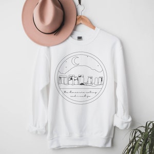 Puede incluir: Sudadera blanca con un dibujo lineal negro de Stonehenge dentro de un c&iacute;rculo, con una luna creciente y estrellas. El texto dice "the stones are calling and i must go". Un sombrero rosa cuelga encima.
