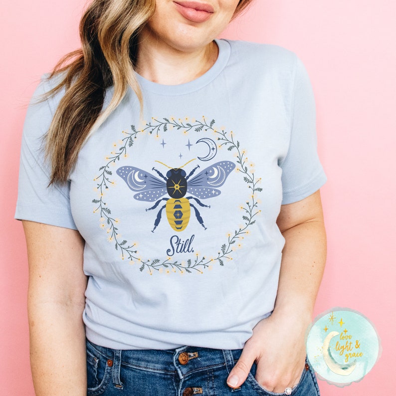 May include: Light blue t-shirt with a graphic of a bee with a moon and stars inside a floral wreath. The text "Still." is below the bee.