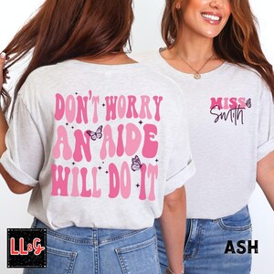 Peut inclure: T-shirt gris clair avec l'inscription rose "DON'T WORRY AN AIDE WILL DO IT" et des papillons. Le devant du t-shirt porte l'inscription "MISS Smith" en rose avec un papillon. Le t-shirt est de couleur cendre.