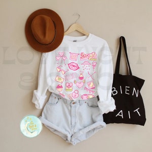 May include: White sweatshirt with a pink and white Valentine's Day themed design. The design includes hearts, arrows, lips, and the words "XOXO".