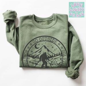 May include: Green sweatshirt with a black graphic of a mountain, a crescent moon, stars, a tent, and a Bigfoot figure. The graphic is in a circle with the text "Go Outside It's Still Worse In Here".