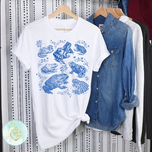 May include: White t-shirt with a blue frog print. The frogs are arranged in a cluster with leaves and vines.