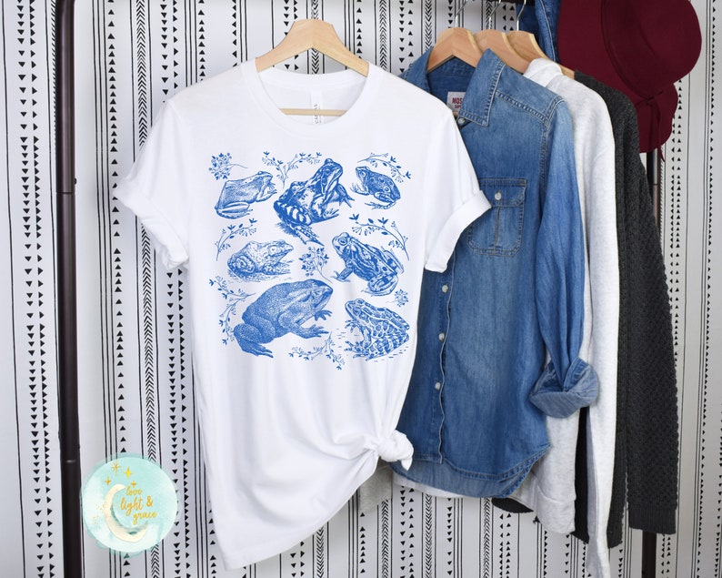 May include: White t-shirt with a blue frog print. The frogs are arranged in a cluster with leaves and vines.