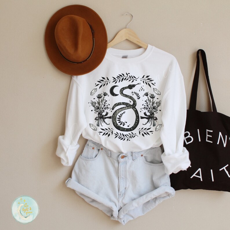 May include: White sweatshirt with a black and white graphic print of a snake with a crescent moon and floral accents. The snake is curled around the moon and has a floral design around it.
