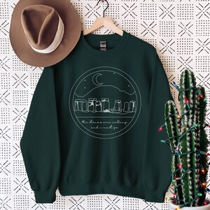 May include: A dark green sweatshirt with a white graphic of Stonehenge, a crescent moon, and stars. The text "the stones are calling and i must go" is below the graphic.