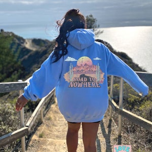 May include: A light blue hoodie with a graphic print on the back that says "Road to Nowhere". The graphic features a desert landscape with a sunset, cacti, and a road leading into the distance. The hoodie is worn by a person standing on a wooden staircase overlooking a body of water.