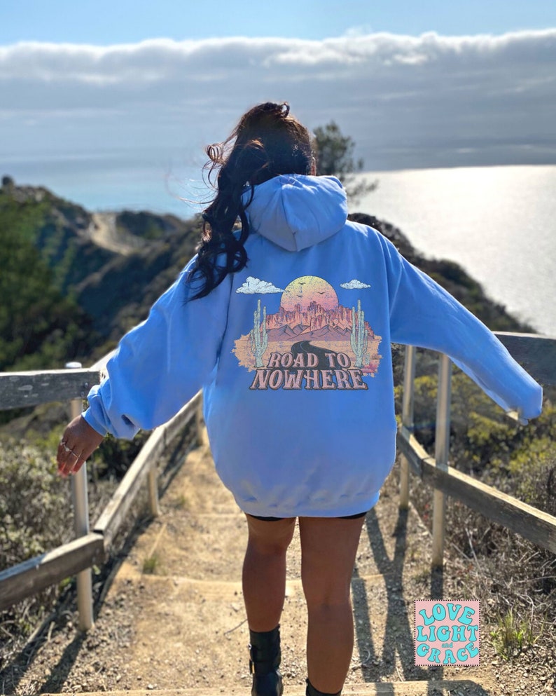May include: A light blue hoodie with a graphic print on the back that says "Road to Nowhere". The graphic features a desert landscape with a sunset, cacti, and a road leading into the distance. The hoodie is worn by a person standing on a wooden staircase overlooking a body of water.