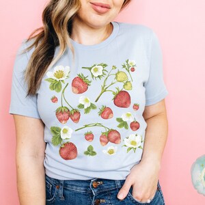 May include: Light blue t-shirt featuring a watercolor design of strawberries, white flowers, and green leaves. The strawberries are red and the flowers are white. The shirt is a soft, comfortable material.