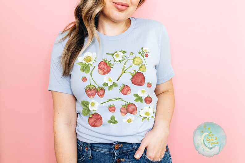 May include: Light blue t-shirt featuring a watercolor design of strawberries, white flowers, and green leaves. The strawberries are red and the flowers are white. The shirt is a soft, comfortable material.