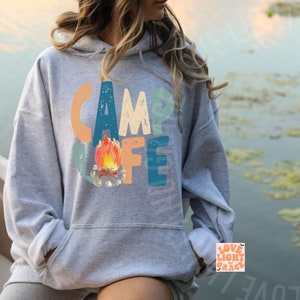 May include: A gray hooded sweatshirt with the words "CAMP LIFE" in colorful letters. A watercolor illustration of a campfire is in the center. The sweatshirt has a front pocket and a drawstring hood.