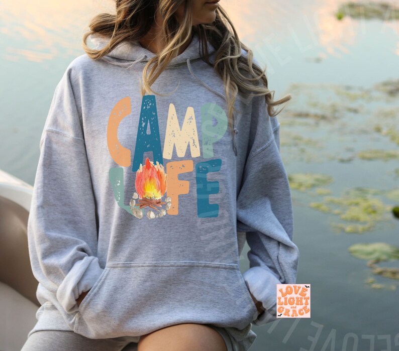 May include: A gray hooded sweatshirt with the words "CAMP LIFE" in colorful letters. A watercolor illustration of a campfire is in the center. The sweatshirt has a front pocket and a drawstring hood.