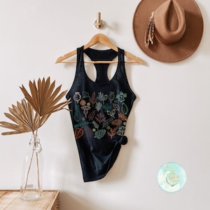 May include: Black racerback tank top with a botanical design featuring various leaves and floral elements in shades of green, orange, and teal. The tank top is hanging on a wooden hanger against a white wall.