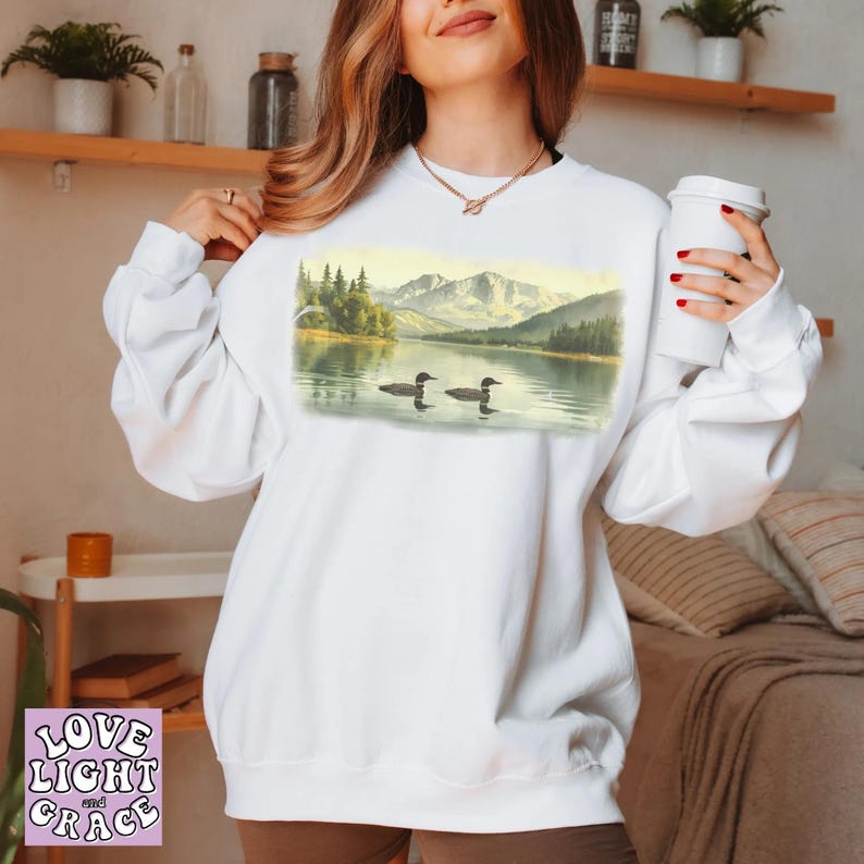 May include: White sweatshirt featuring a scenic landscape with a lake, mountains, and two loons. The graphic is a watercolor painting style. The sweatshirt has long sleeves and a crew neck.