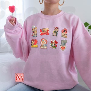 May include: A pink sweatshirt with a vintage Valentine's Day theme. The sweatshirt features nine different images of couples and animals with hearts and romantic messages. The text "Love Light & Grace" is printed on the bottom left corner of the sweatshirt.