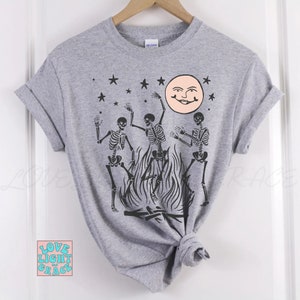 May include: A gray t-shirt with a graphic of three skeletons dancing around a bonfire with a smiling moon and stars in the background.