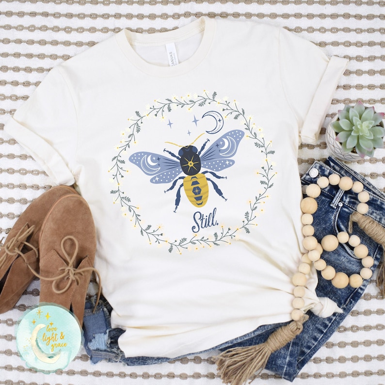 May include: A white t-shirt with a blue and yellow bee illustration surrounded by a floral wreath. The bee has a crescent moon on its wings and the word "Still" is written below it.