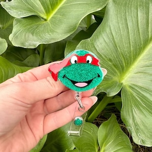 May include: Green and red glitter badge holder featuring a cartoon Teenage Mutant Ninja Turtle character with a red bandana. The holder has a clear plastic retractable badge clip.