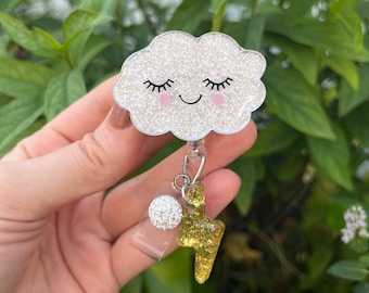 May include: A white glitter cloud with a smiling face and closed eyes, attached to a retractable badge holder with a sparkly silver charm and a gold glitter lightning bolt charm.