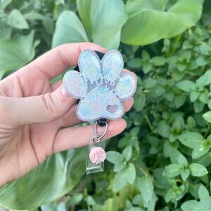 May include: A white and iridescent glitter paw print with the text "Mrs. Barton" and a small pink heart. The paw print is attached to a black retractable badge holder with a pink glitter charm.