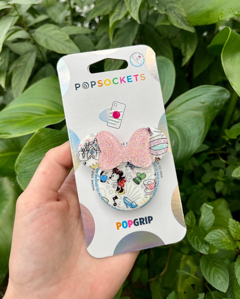 May include: A pink and white PopSocket phone grip with a Minnie Mouse design. The design features Minnie Mouse in a red dress and white polka dots, a castle, and a teacup. The text on the PopSocket reads 'Let's Go To Disney World' and 'Phone Grip & Telephone Stand'. The PopSocket is packaged on a white card with the PopSockets logo and the word 'PopGrip'.