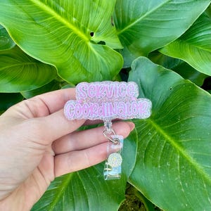 May include: A pink and silver glitter badge reel with the words "Service Coordinator" in a script font. The badge reel has a clear plastic holder and a decorative silver accent.