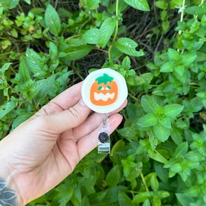 May include: A white pumpkin-shaped retractable badge holder with a black and green jack-o'-lantern face. The holder is attached to a black and silver retractable badge reel.