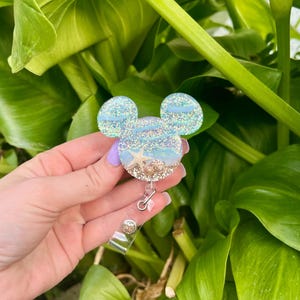 May include: A blue and white glitter resin Mickey Mouse head with a seashell and starfish design. The head is attached to a retractable badge holder.