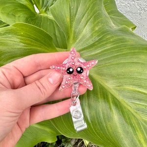 May include: Pink glitter starfish with a cute cartoon face and pearl accents. The starfish is attached to a retractable badge reel.