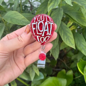 Könnte beinhalten: Roter Glitzerballon mit der weißen Aufschrift "You'll Float Too", befestigt an einem durchsichtigen Plastik-Badgehalter mit einem roten Glitzerkreis.