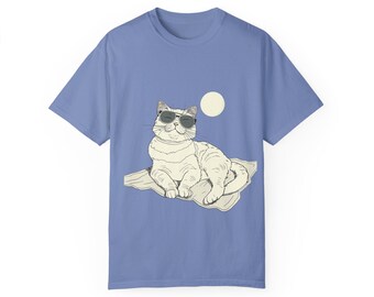 May include: A light blue t-shirt featuring a graphic of a cat wearing sunglasses and lying on a beach towel. The cat is white with black markings and has a relaxed expression. The sun is shining in the background.