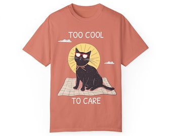 May include: A coral colored t-shirt with a black cat wearing sunglasses and a bow tie sitting on a picnic blanket. The text "TOO COOL TO CARE" is printed on the shirt.
