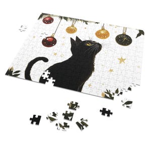 May include: A black cat sits on a white background with a Christmas tree in the background. The cat is looking up at a Christmas ornament hanging from the tree. The ornament is gold and has a star on it. There are other ornaments hanging from the tree, including red and gold ornaments.