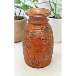 May include: A rustic, orange-colored wooden vase with a distressed finish. The vase has a wide, rounded body and a narrow neck with a flared rim. The surface shows visible wood grain and dark brown accents, suggesting an aged appearance. Decorative home decor.