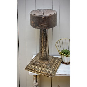 Puede incluir: Un pedestal de metal antiguo con base cuadrada y parte superior cilíndrica. El pedestal tiene un diseño ornamentado y texturizado y un acabado envejecido similar al bronce. Una pequeña planta suculenta en una cesta de alambre dorado está cerca.