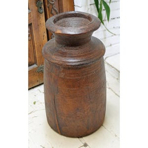 May include: A tall, cylindrical wooden vessel with a dark brown, weathered finish. The object features a wide, rounded rim, a distinct neck section, and a straight body with visible wood grain texture and minor surface wear throughout the exterior.