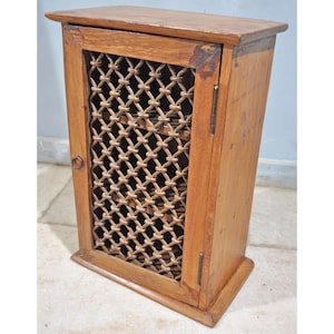 May include: A small wooden cabinet with a single door. The door is made of a woven metal grate and has a small metal ring handle. The cabinet is a light brown colour.