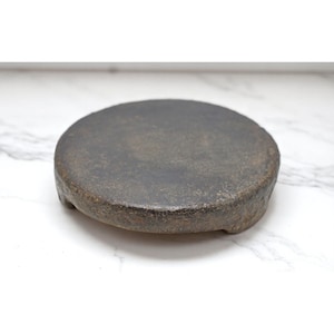 Puede incluir: Un mortero de piedra redondo, de color gris oscuro, con un borde ligeramente elevado y tres pequeños pies. El mortero tiene una superficie rugosa y texturizada y parece estar hecho de un material pesado. Se encuentra sobre un fondo de mármol blanco.