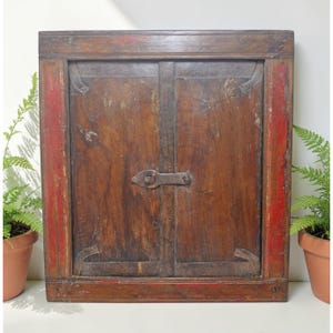 May include: A vintage wooden cabinet with two doors, finished in a dark brown hue with visible wood grain. The frame has red accents on the sides. It is secured with a metal latch and hinges, displaying an antique aesthetic.