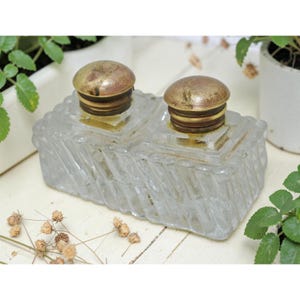 May include: A vintage clear glass inkwell with two wells, topped with brass-colored lids. The inkwell has a textured, geometric design on the sides. The lids show signs of age, adding to the antique aesthetic. A classic desk accessory.
