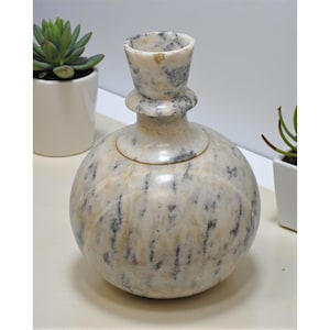 May include: A decorative marble vase with a round body and a flared top. The vase is primarily off-white with gray and black veining. The vase is next to two small potted plants.
