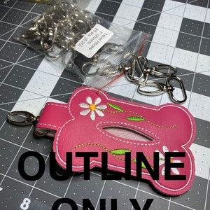 Puede incluir: Porta pañuelos de tela rosa con un diseño de margarita y bordados verdes. El porta pañuelos tiene un lazo de metal para sujetarlo a una bolsa o bolso. El texto "OUTLINE ONLY" está impreso en una superficie negra debajo del porta pañuelos.
