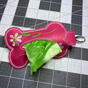 May include: A hot pink dog waste bag dispenser shaped like a bone, featuring white stitched edges and an embroidered white daisy with a yellow centre and green stem. A bright green plastic bag protrudes from the centre, and a silver metal clasp is attached to the side.